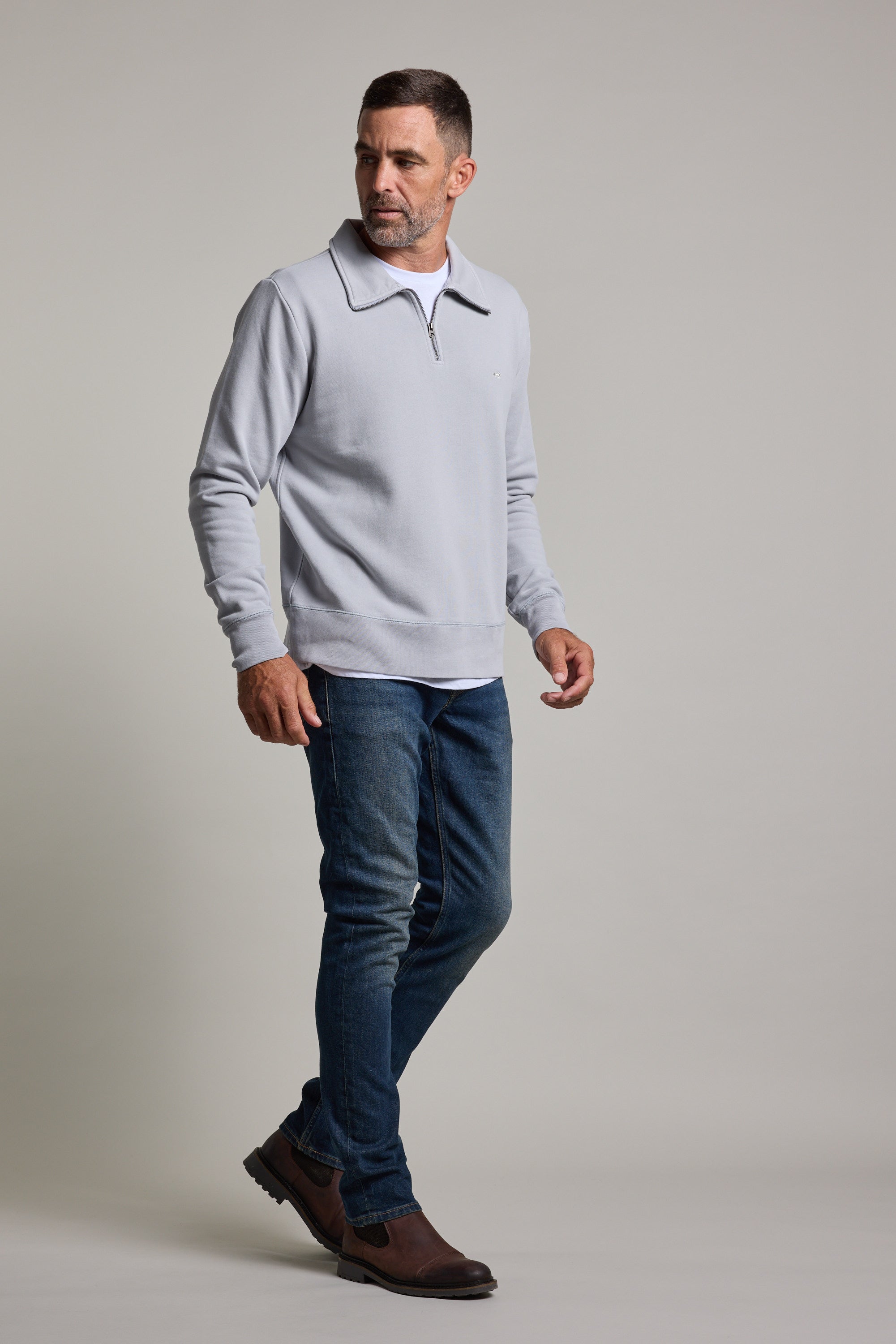A man with short dark hair and a trimmed beard stands against a plain light background, wearing the Barkers Galway GD 1/4 Zip, blue jeans, and brown shoes.