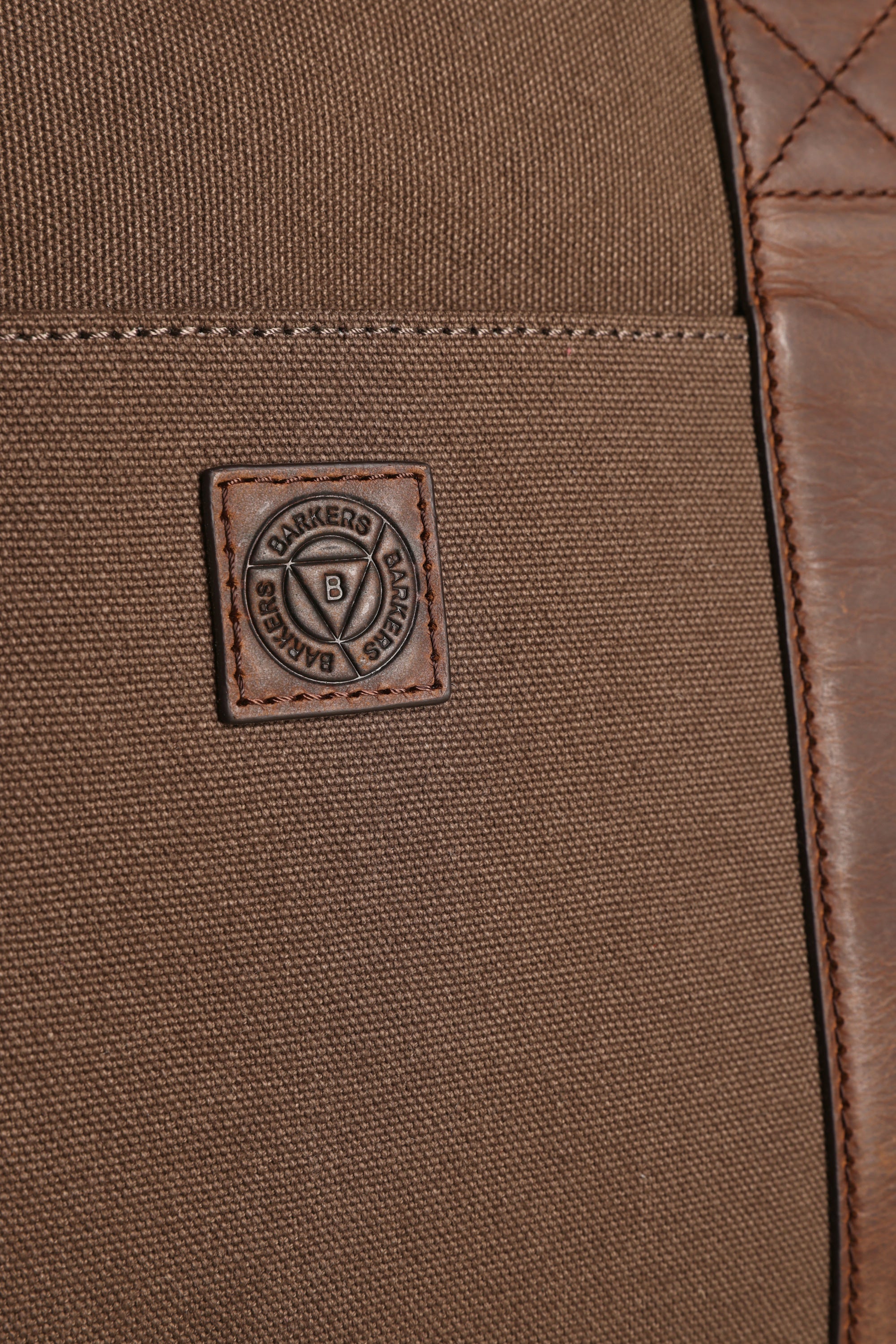 Close-up of the Newman Canvas Weekender, a brown canvas and leather travel bag by Barkers, featuring a stitched rectangular Barkers brand patch with a circular emblem in the center.