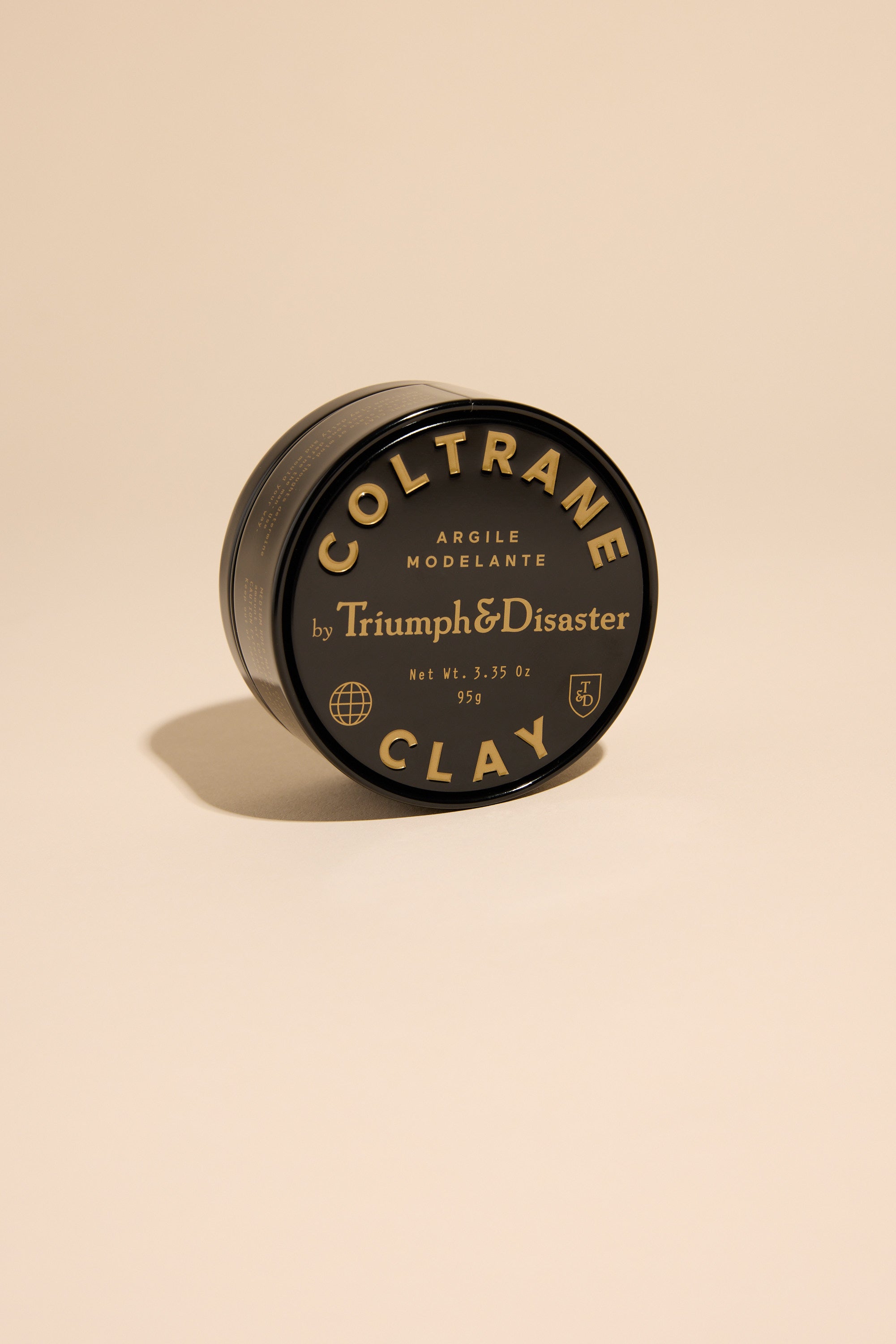 A black tin of Triumph & Disaster Coltrane Clay by Triumph & Disaster, featuring gold and white label text, is displayed on a plain beige background with the round container slightly tilted.