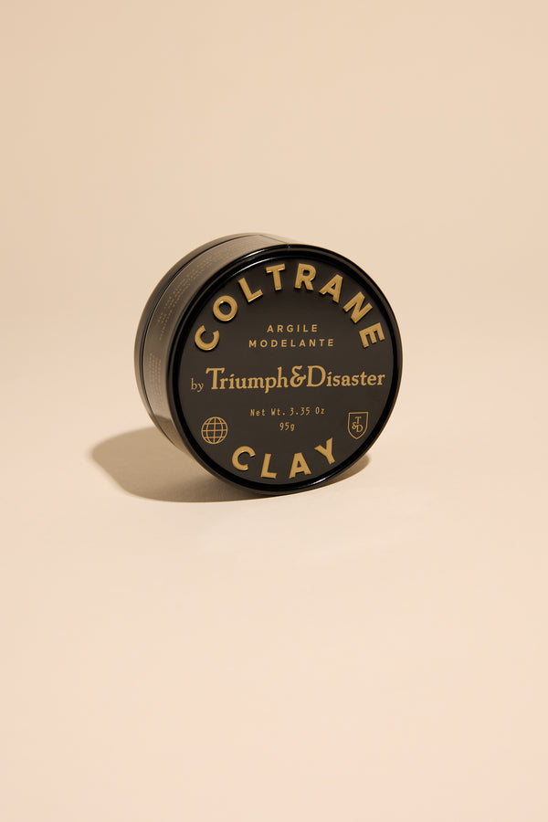 A black tin of Triumph & Disaster Coltrane Clay by Triumph & Disaster, featuring gold and white label text, is displayed on a plain beige background with the round container slightly tilted.