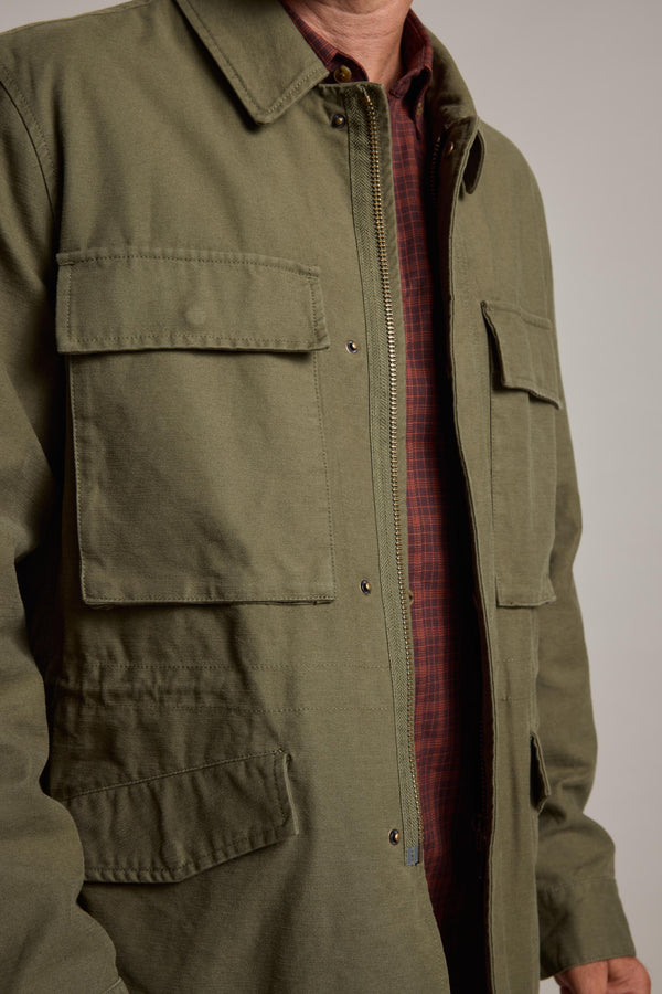 A person wears the Barkers Nowell Field Jacket in olive green cotton with large front pockets over a red and gray plaid shirt, shown from shoulders to waist. The face is not visible, emphasizing classic utilitarian style.