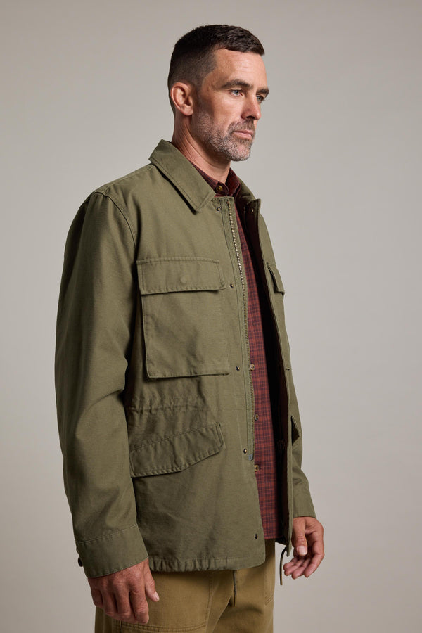 A man with short dark hair and a beard wears the Barkers Nowell Field Jacket in olive green over a brown plaid shirt and tan pants, standing against a plain light background.