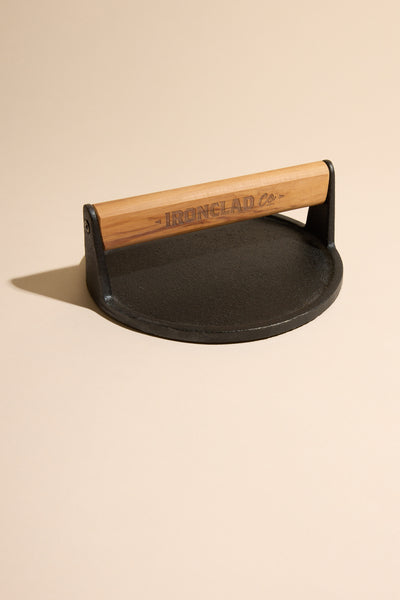 The IronClad Burger Press—a black cast iron press with a wooden handle engraved with IronClad—rests on a beige surface, casting a subtle shadow. Perfect for crafting delicious smash burgers.
