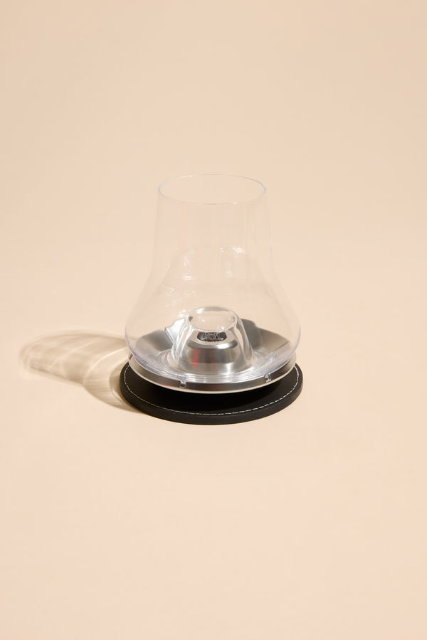 The Other Whisky Glass Individual, a clear stemless nosing glass with a wide base and narrow top, rests on a black circular coaster against a beige background, casting a soft shadow to the left.