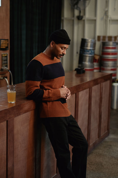 Wearing the Barkers Portway Stripe Merino Crew and a black beanie, a man leans on a wooden bar with a glass of beer. He smiles while looking down, with beer kegs and dark curtains behind him.