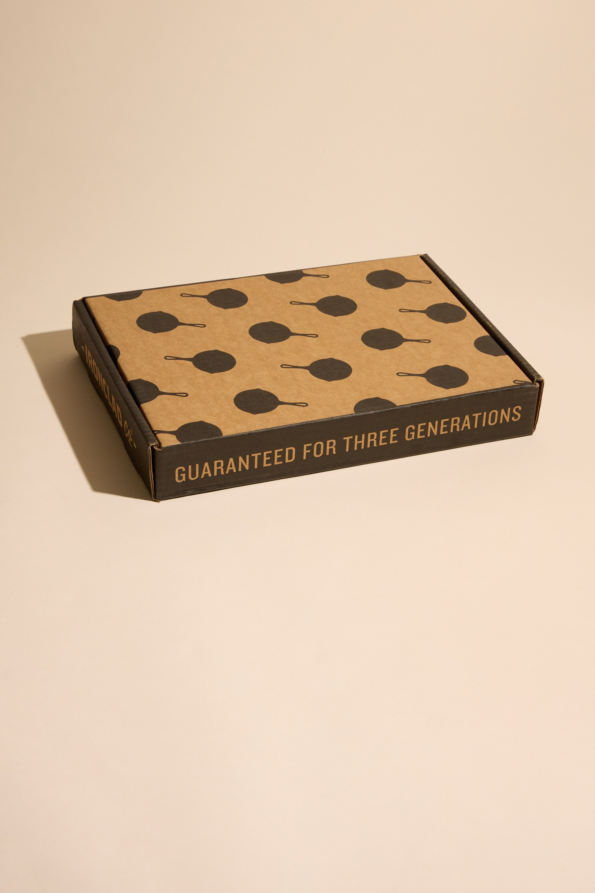 A flat cardboard box featuring dark skillet silhouettes on the lid and “GUARANTEED FOR THREE GENERATIONS” on the side holds the Other Ironclad Lil' Legacy Pan, a compact cast iron or induction-compatible skillet. The box sits on a beige surface.