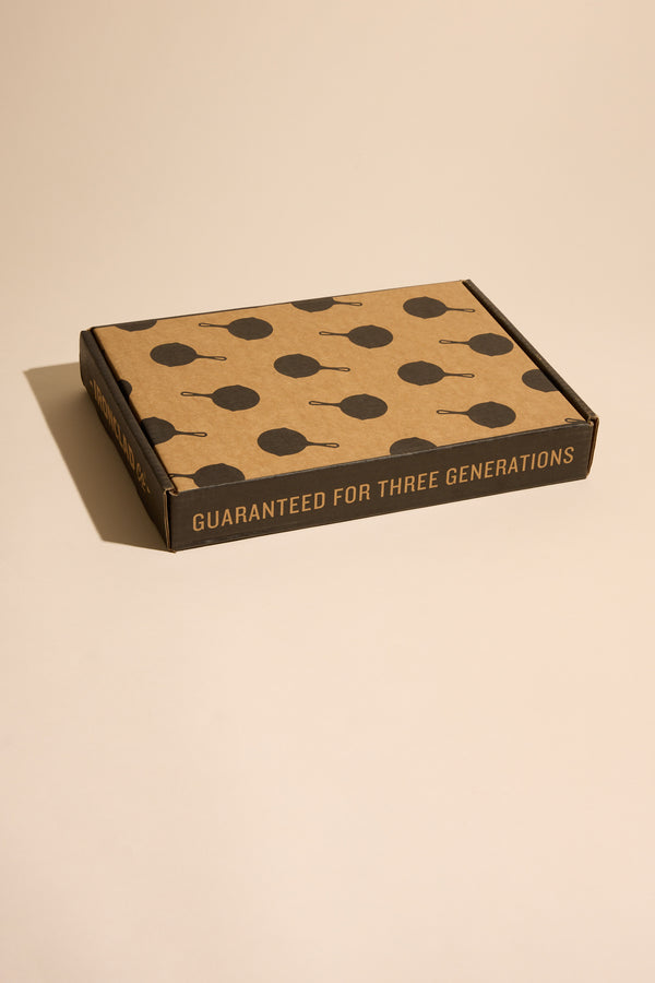 A flat cardboard box featuring dark skillet silhouettes on the lid and “GUARANTEED FOR THREE GENERATIONS” on the side holds the Other Ironclad Lil' Legacy Pan, a compact cast iron or induction-compatible skillet. The box sits on a beige surface.