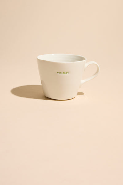 The Bucket Mug - Old Fart by Other is a white ceramic mug featuring "old fart" in small green lettering, set on a beige surface with a soft shadow—an ideal humorous gift for an old fogey.