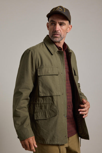 A man wears the Barkers Nowell Field Jacket in olive green over a brown plaid shirt and khaki pants, paired with a dark baseball cap, as he looks to his left against a plain light background.