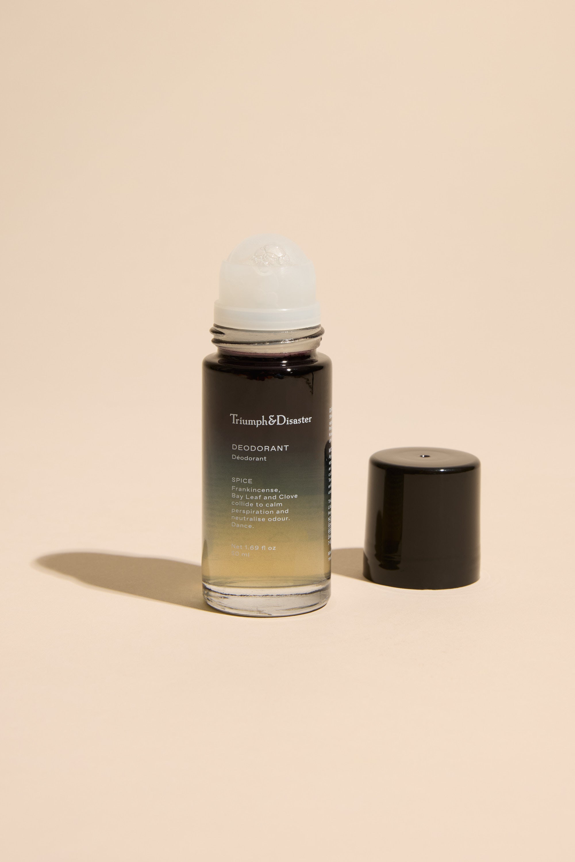 A Triumph & Disaster Deodorant V1 Spice bottle stands upright on a beige surface, its black cap off and beside it. The organic roll-on shows a gradient liquid inside, from clear to dark.