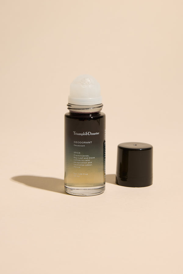 A Triumph & Disaster Deodorant V1 Spice bottle stands upright on a beige surface, its black cap off and beside it. The organic roll-on shows a gradient liquid inside, from clear to dark.