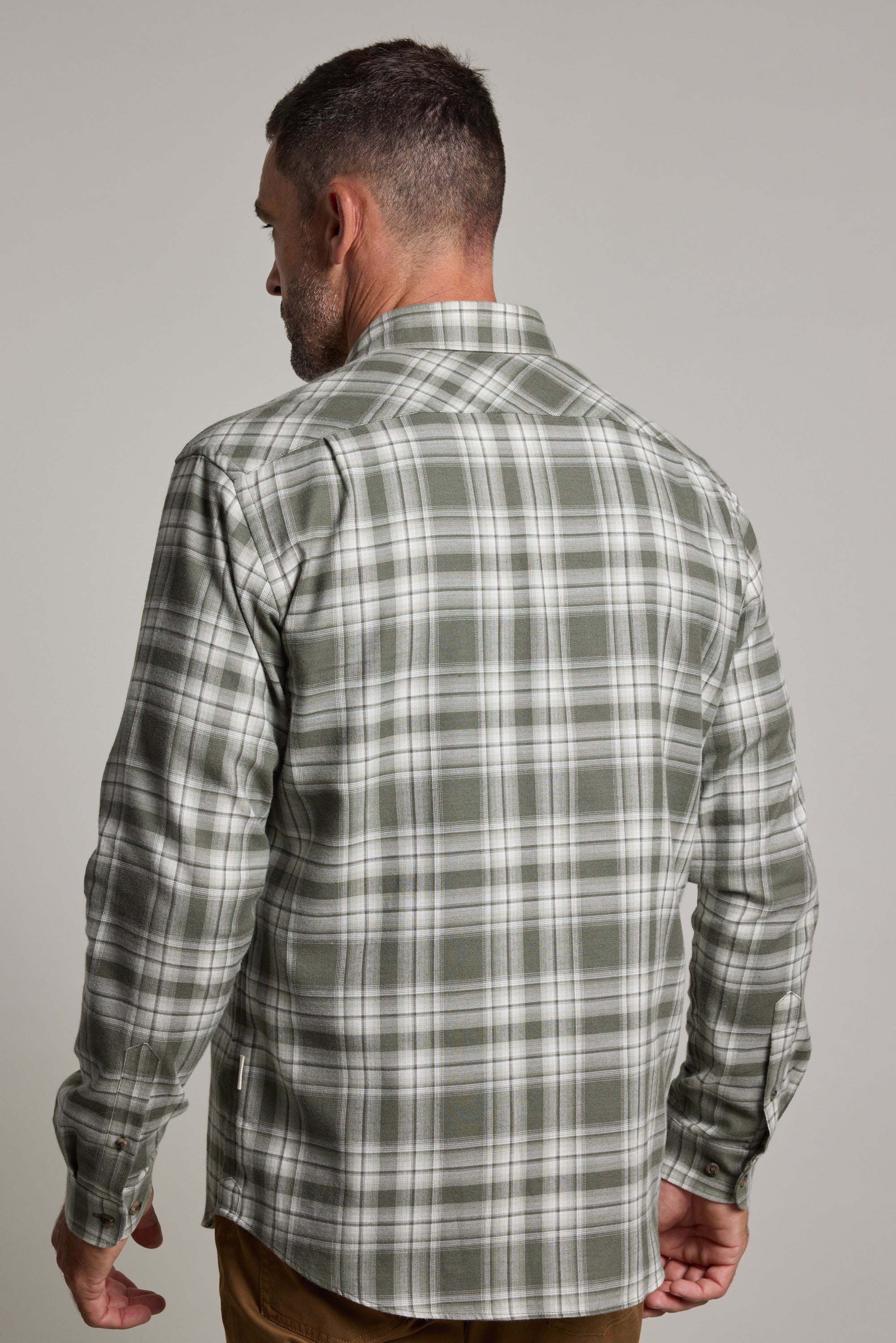 A man stands with his back to the camera, wearing a green check Barkers Paxtonheck Shirt in a classic fit and brown pants, set against a plain light gray background.