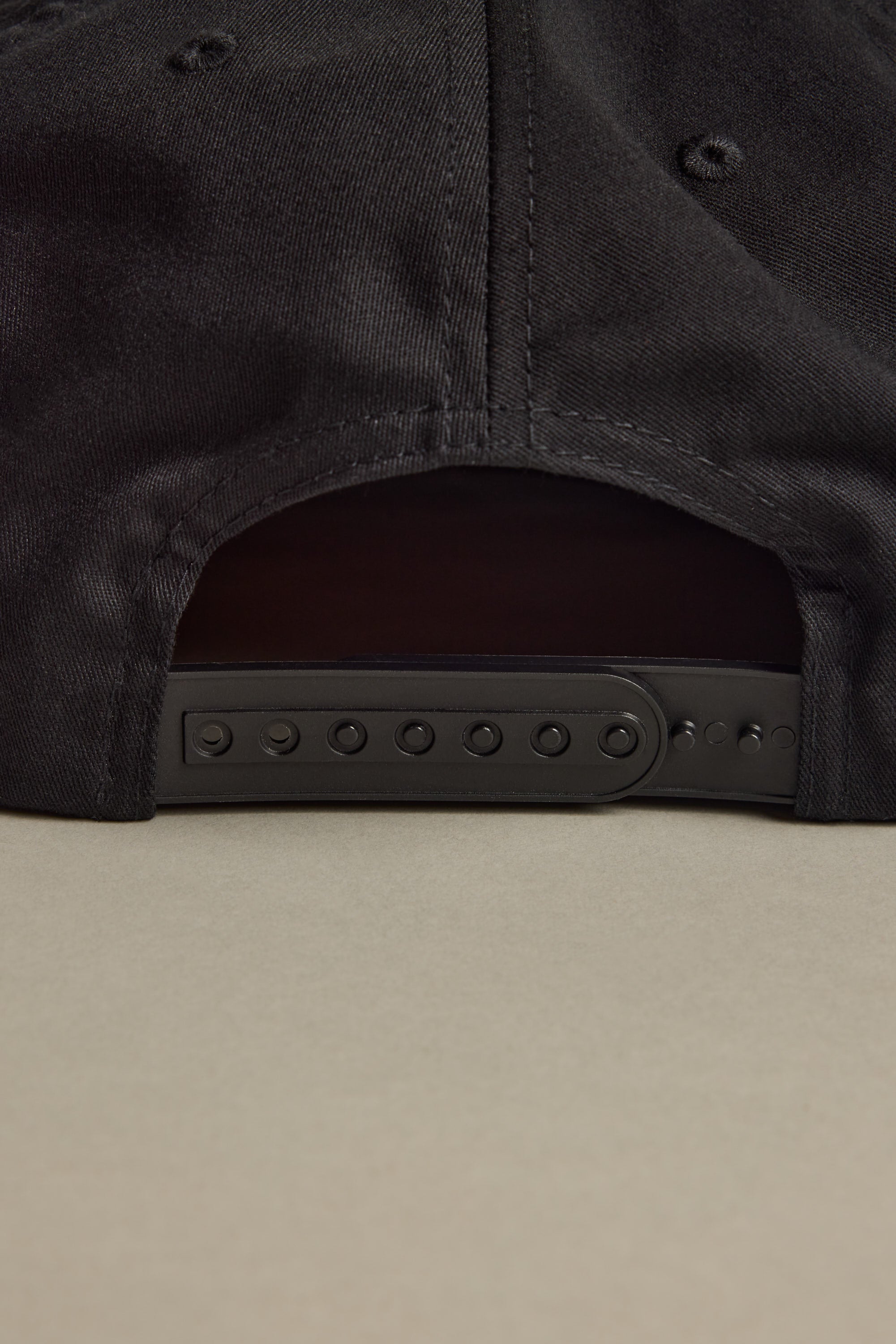 Close-up of the back of the Brixton Payment MP Snapback by Brixton, showing its black unstructured 5-panel design and adjustable plastic snap closure, resting on a light beige surface.