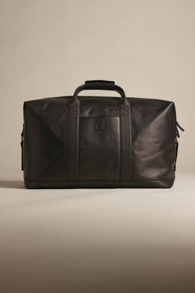 The Barkers Costello Leather Weekender, a spacious black duffel bag with dual handles and a shoulder strap, is displayed on a plain beige background.