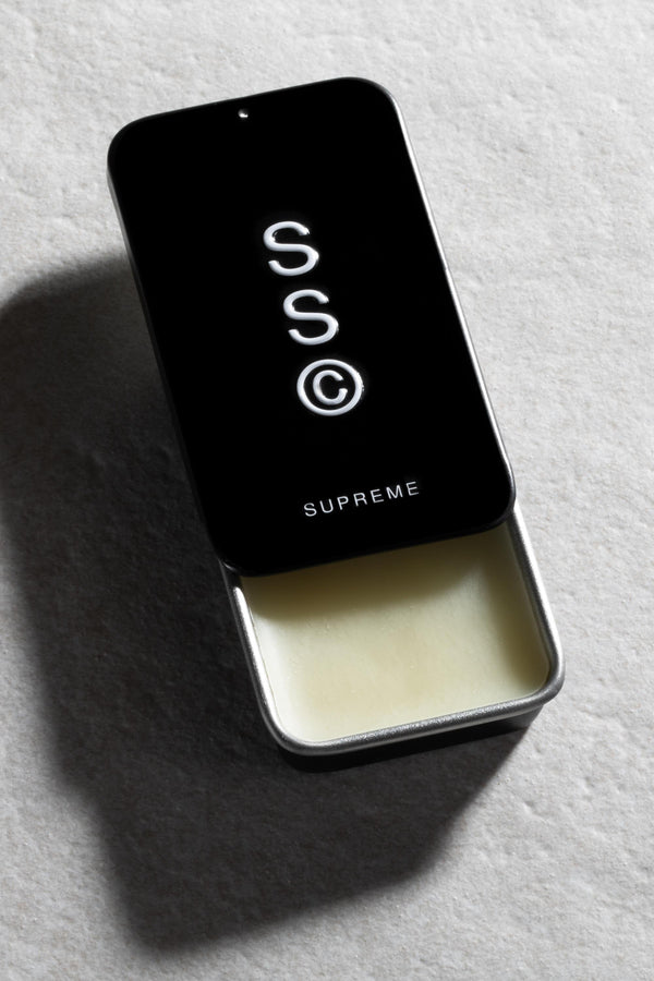 A partially open rectangular metal tin with a black sliding lid labeled "Solid State - Supreme" reveals pale wax cologne inside. The Solid State tin rests on a textured light gray surface.