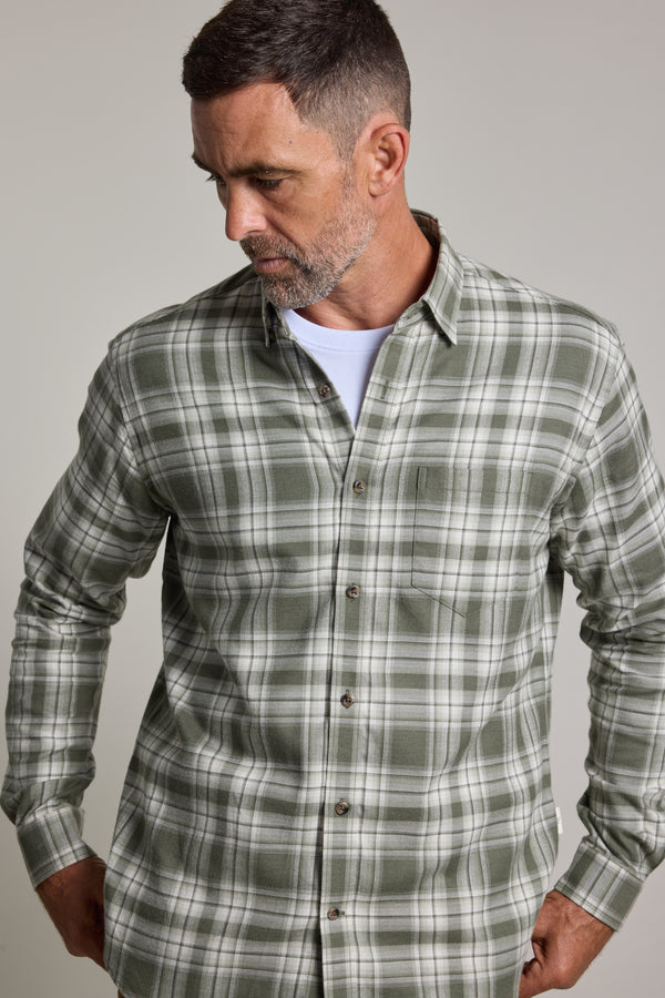 A man with short dark hair and a trimmed beard wears the Barkers Paxtonheck Shirt in green check over a white T-shirt, looking down and to the side with his hands in his pockets.