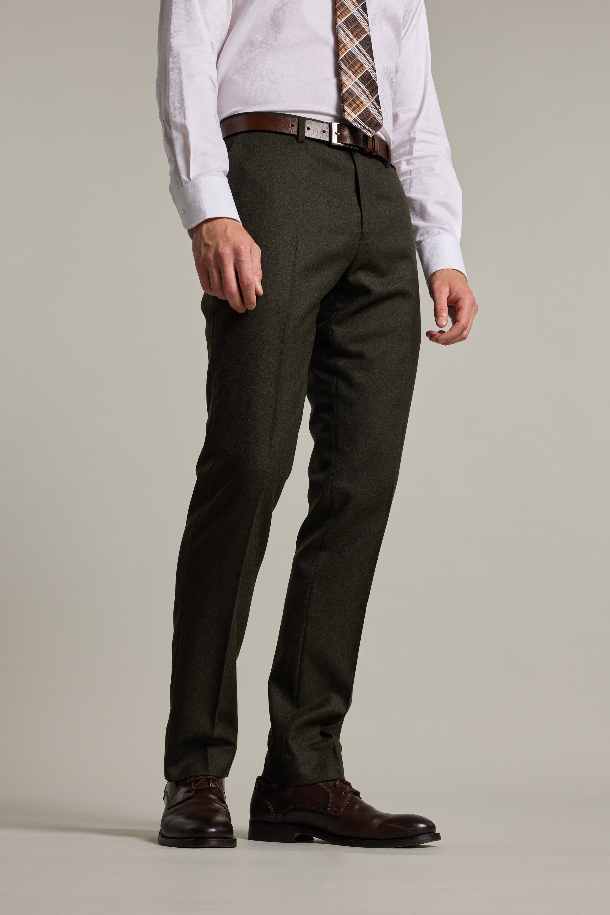 A man wearing Barkers Howard Suit Trouser in forest green, paired with a white shirt, brown belt, plaid tie, and brown dress shoes stands against a plain light background. His face is not visible.