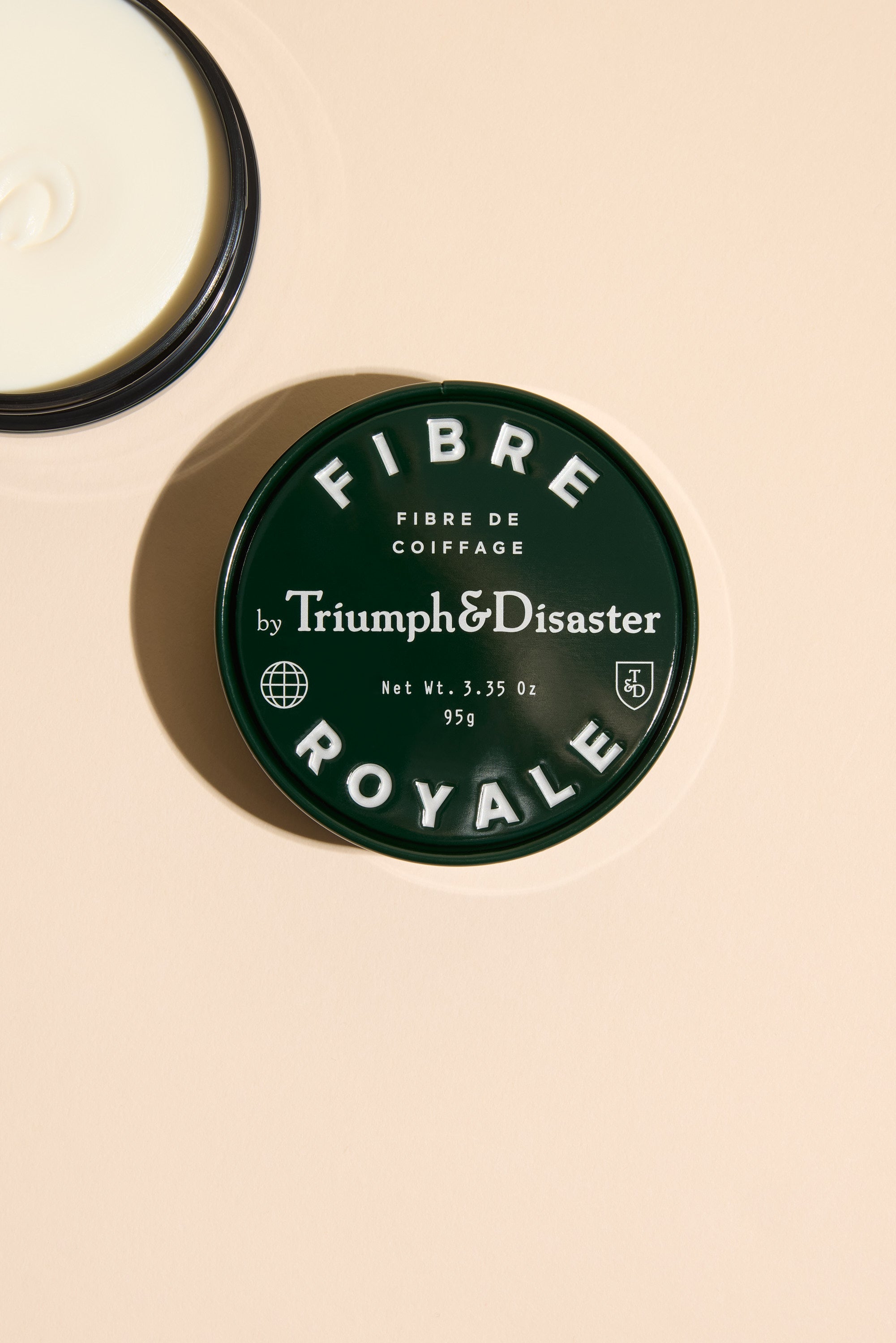 A dark green tin labeled Triumph & Disaster Fibre Royale, a strong hold hair product by Triumph & Disaster, rests on a beige surface beside an open container revealing a swirl of white cream inside.