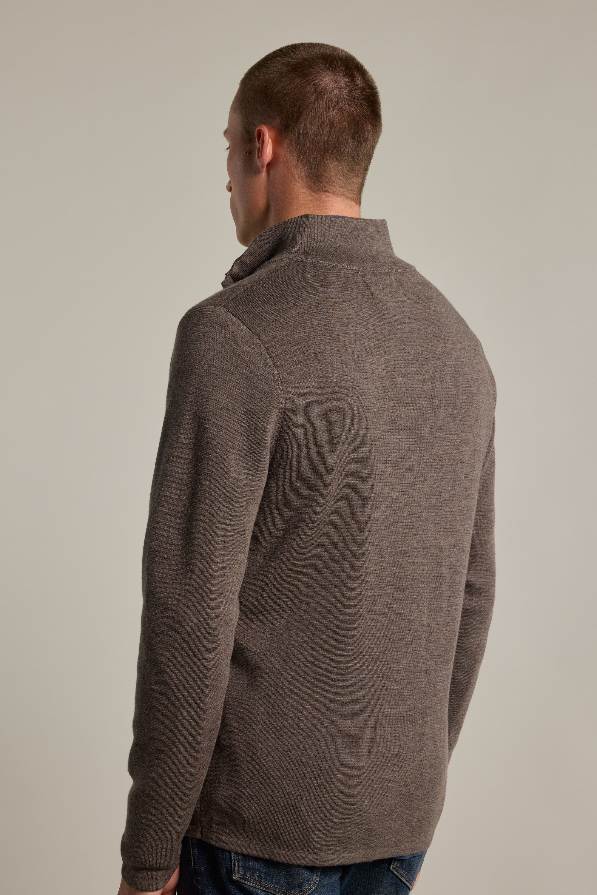 A person wearing the Barkers Brandon ZQRX Merino 1/4 Zip sweater in brown, styled as a layering piece with blue jeans, stands with their back to the camera against a plain light background.
