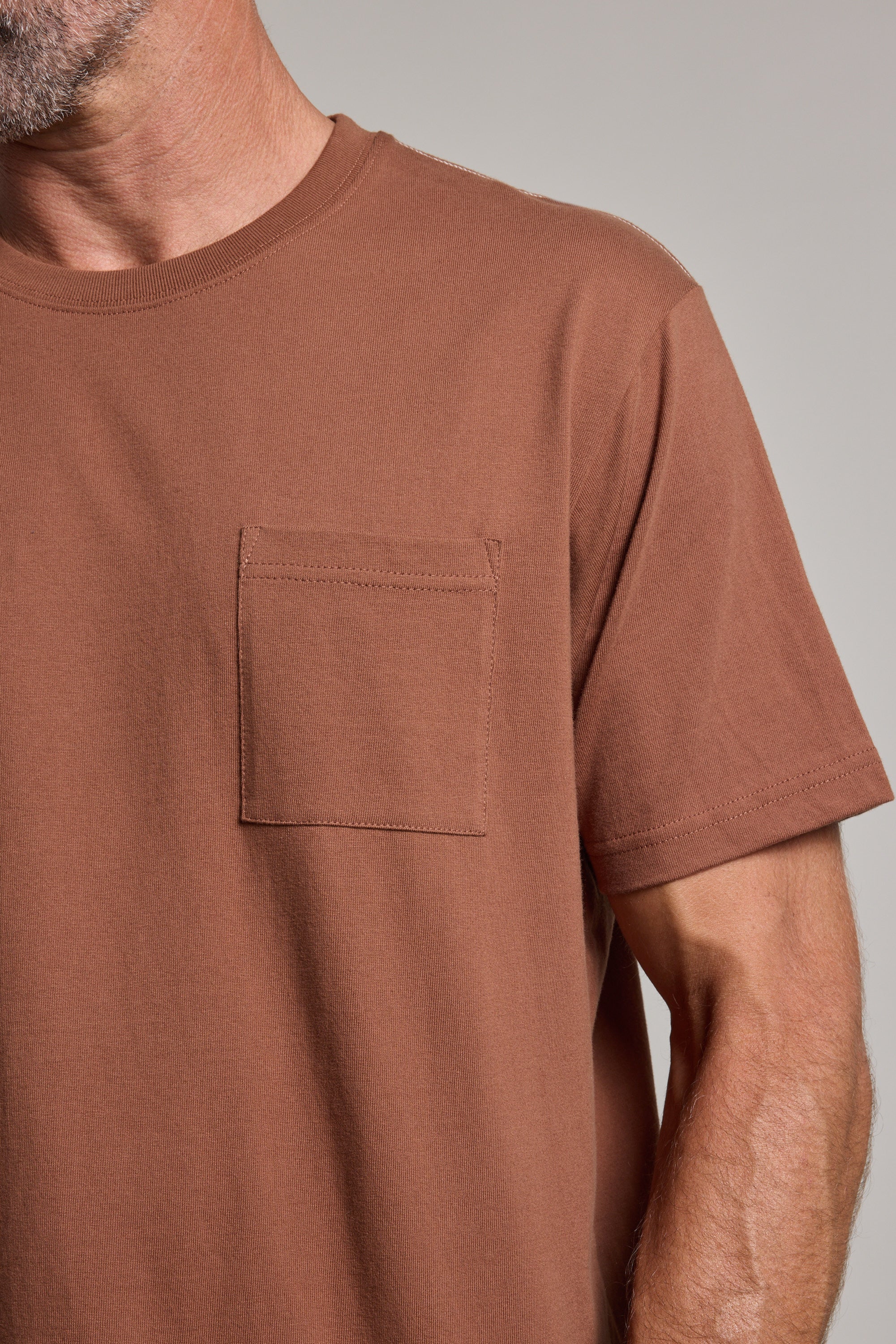 A close-up of a person wearing the Barkers Wrenches Print Tee, a brown heavyweight t-shirt featuring a small chest pocket on the left; only the lower face, neck, and upper body are visible.