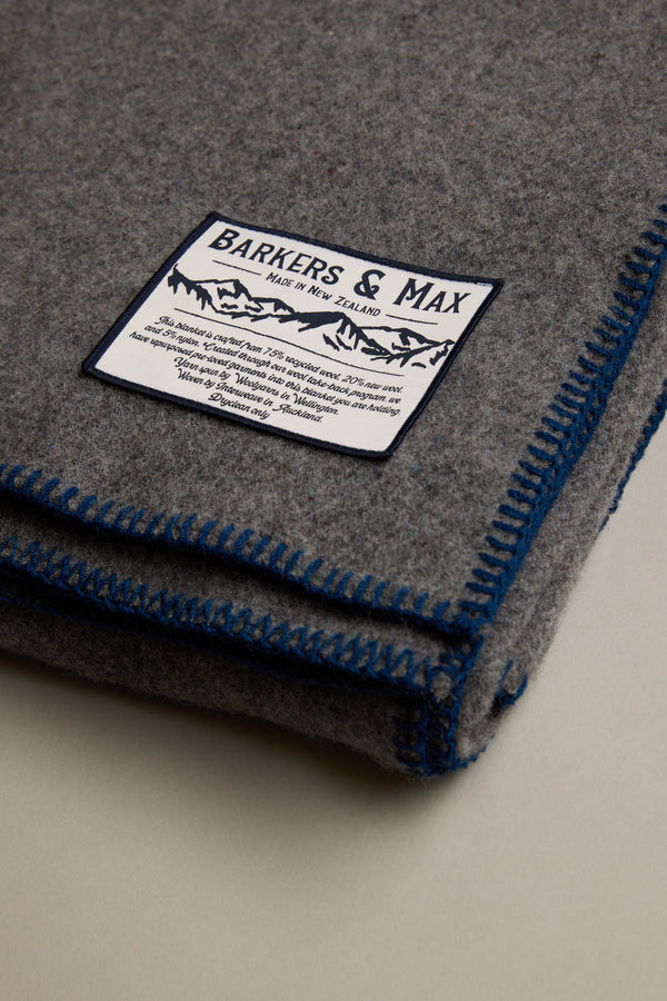 A folded grey Wool Take Back Blanket by Other, made from sustainable wool, features blue-stitched edges and a white label with text and mountain graphics, resting on a light surface.