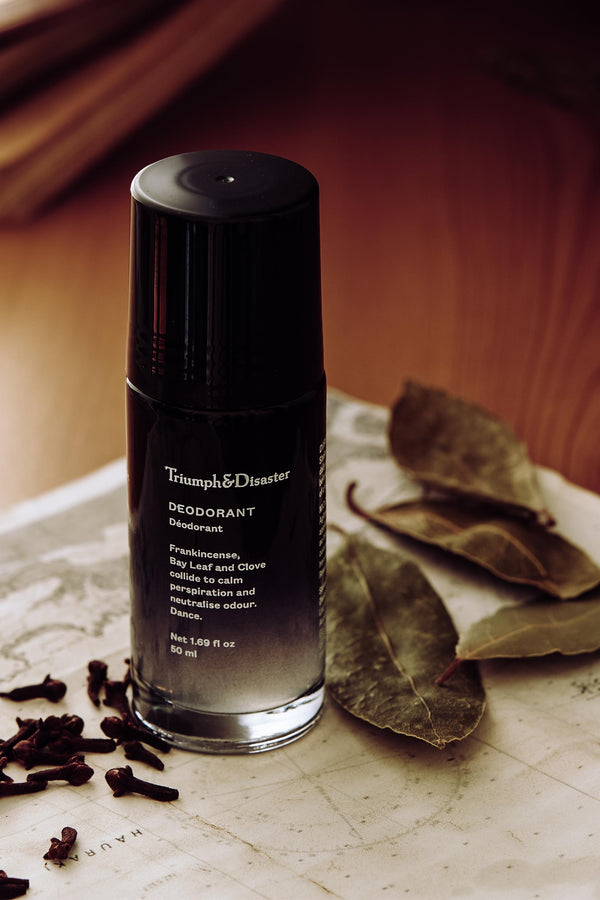 A bottle of Triumph & Disaster Deodorant V1 Spice sits on a map beside dried bay leaves and clove buds, arranged on a wooden surface under warm, soft lighting.