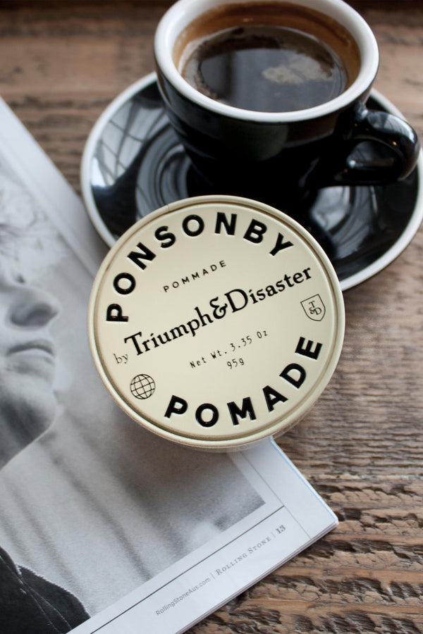 A tin of Triumph & Disaster Ponsonby Pomade, a natural hair and scalp treatment, rests on an open magazine beside a black coffee cup on a wooden table.