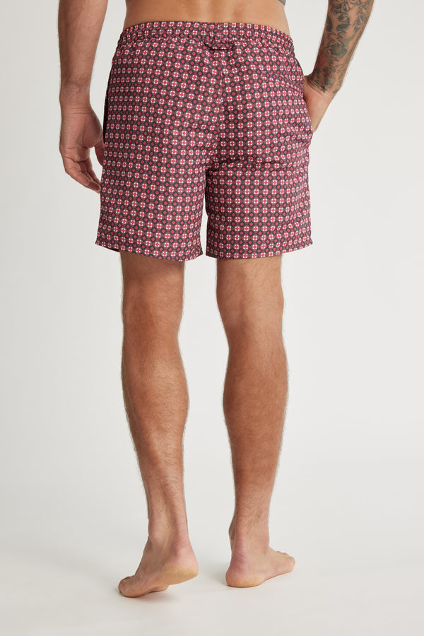 A man stands barefoot facing away, wearing Barkers Laramie Print Volley shorts with a repeating geometric maroon pattern made from recycled polyester. A tattoo is visible on his right arm against a plain white background.