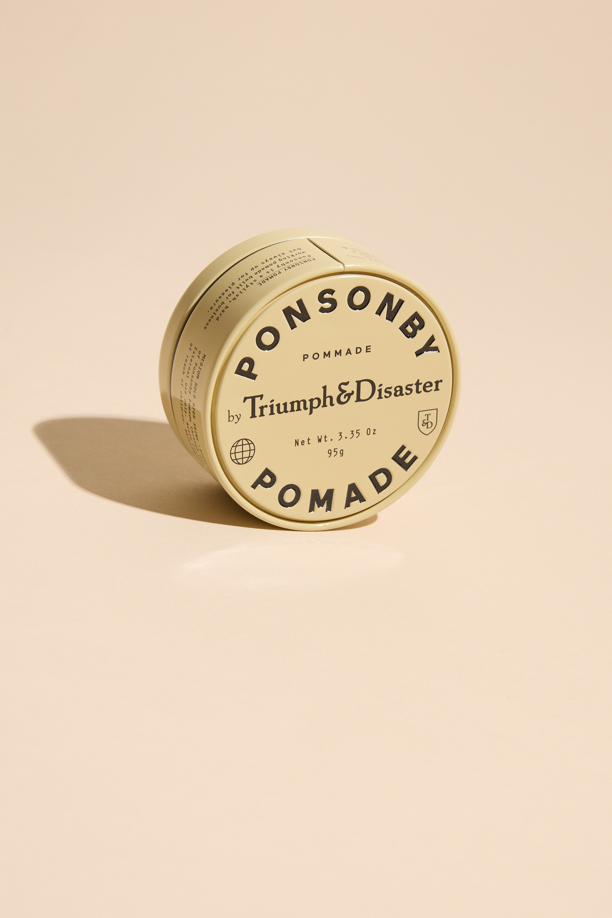 A round tin of Triumph & Disaster Ponsonby Pomade with beige packaging and bold black text rests on a light beige surface, casting a soft shadow.