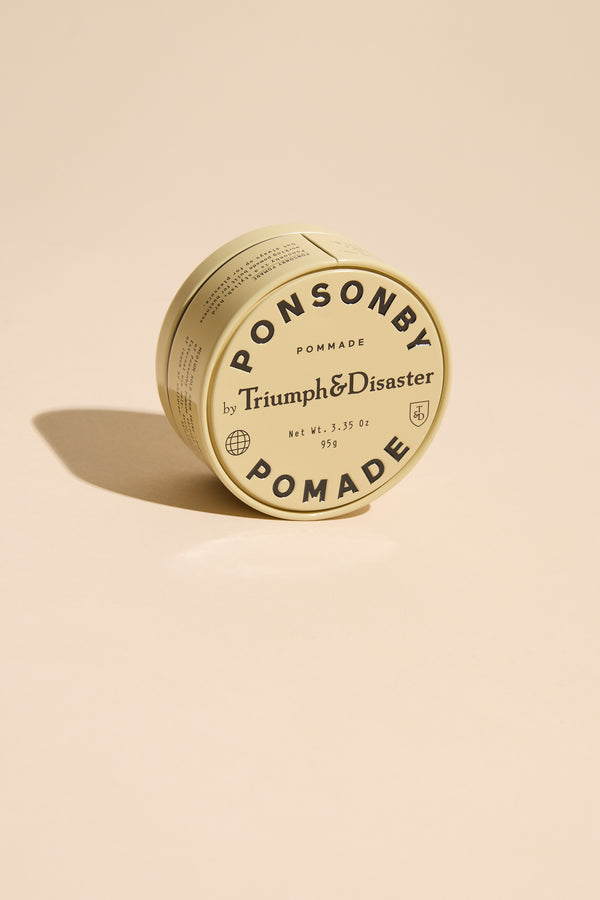 A round tin of Triumph & Disaster Ponsonby Pomade with beige packaging and bold black text rests on a light beige surface, casting a soft shadow.