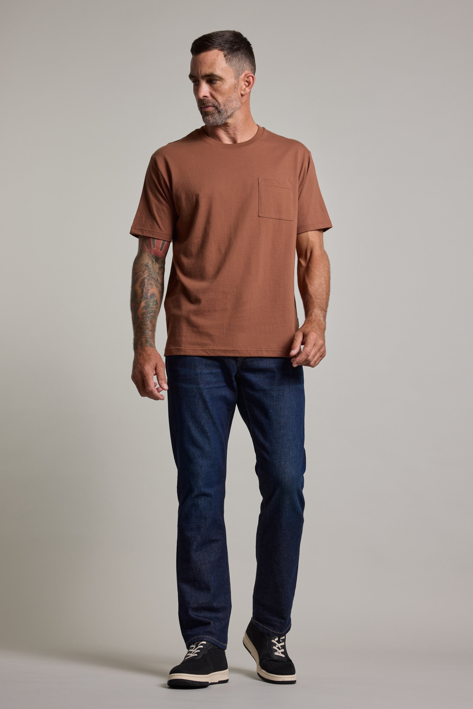 A man with a short beard and a tattooed arm stands slightly sideways against a light gray background, wearing the Barkers Wrenches Print Tee in brown, dark blue jeans, and black sneakers.