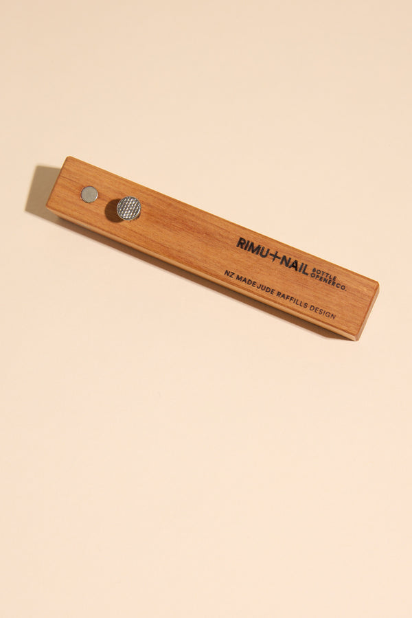 A Rimu & Nail Bottle Opener by Other, engraved with NZ MADE/JUDE RAFFILLS DESIGN, rests on a light beige surface—crafted by the maker behind handmade New Zealand designs.