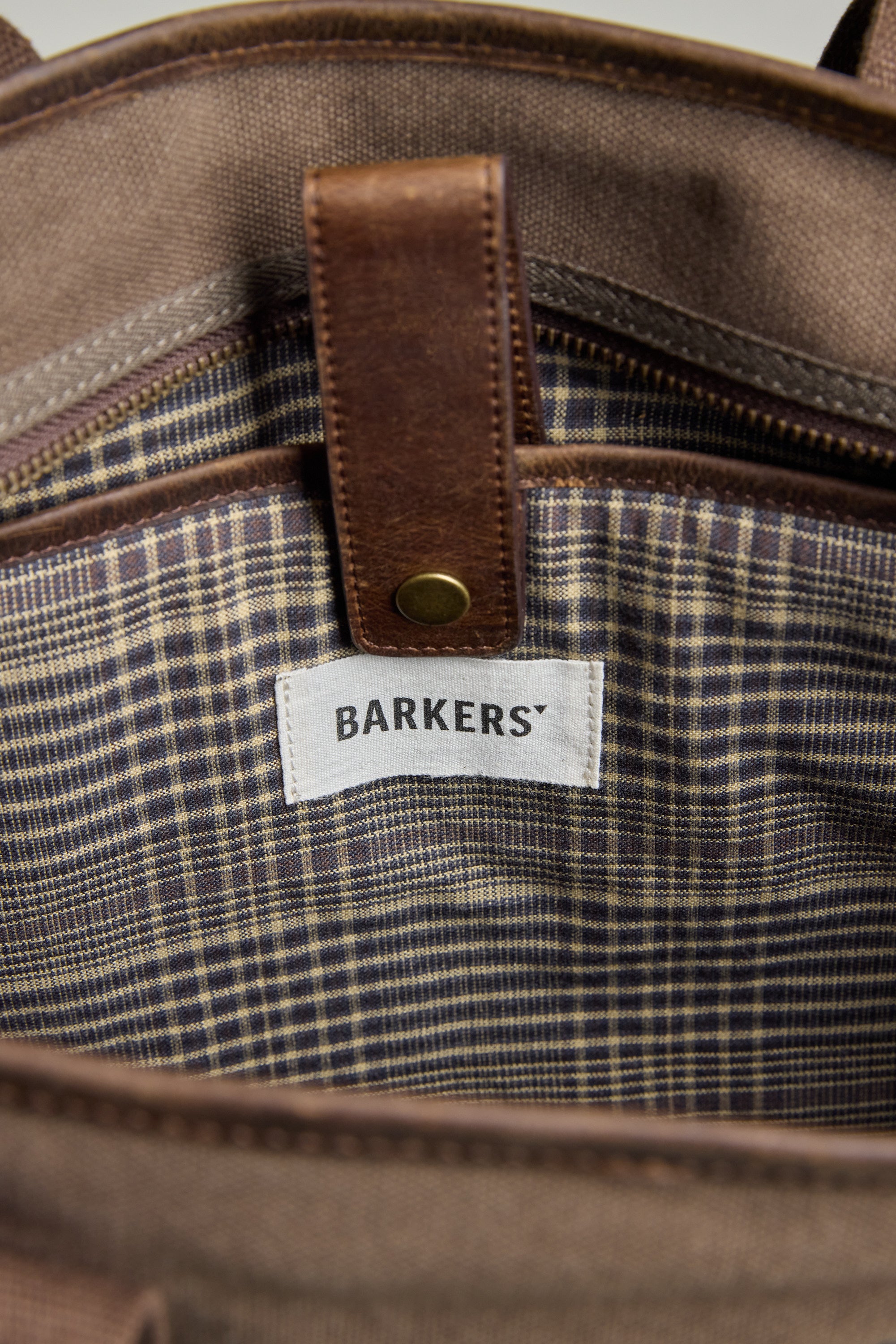 Close-up of the Foxton Canvas Convertible Tote by Barkers, showing its plaid fabric lining and a white Barkers label under a brown leather strap with a brass snap button.