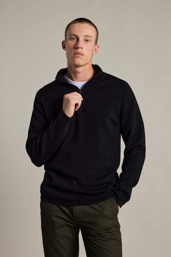 A young man poses against a plain background, wearing the Barkers Brandon ZQRX Merino 1/4 Zip in black over a white shirt and dark green pants. He looks at the camera with a neutral expression, holding the sweater’s collar—ideal for cold-weather layering.