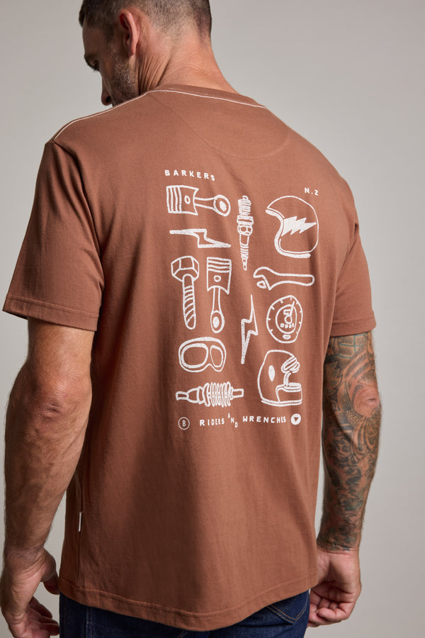 A man wearing the Barkers Wrenches Print Tee, a heavyweight graphic shirt featuring white line drawings of tools and mechanical parts like a wrench, spark plug, and helmet on the back, stands facing away to display the design.