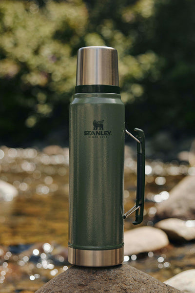 The Stanley Classic Legendary Bottle | 1L by Stanley, featuring a stainless steel lid and handle, rests on a rock near a stream, seamlessly blending with outdoor adventure gear amid blurred greenery and flowing water in the background.