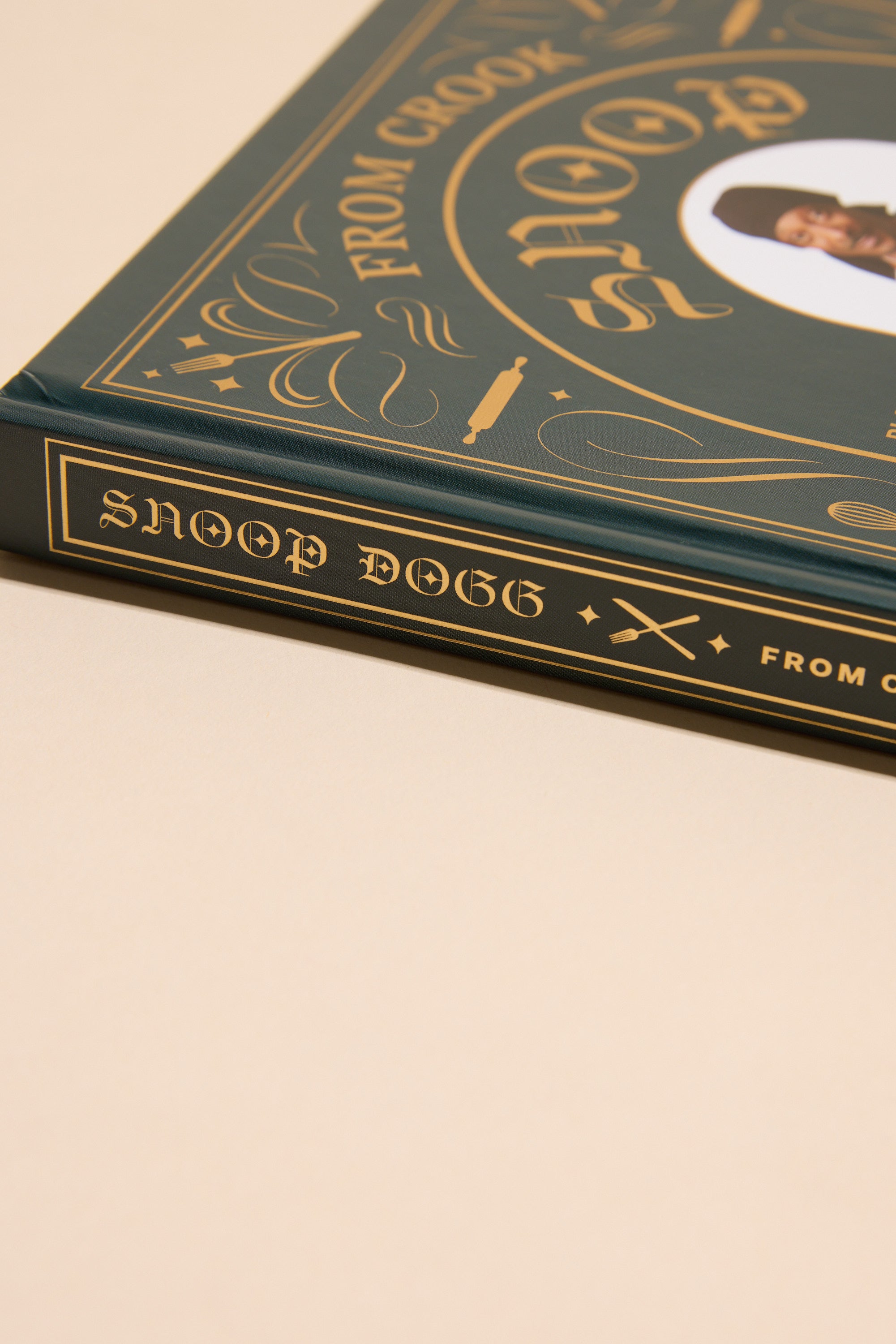 A close-up of the spine and corner of a hardcover cookbook titled "From Crook to Cook" by Other, featuring decorative gold lettering and designs on a dark green cover with cannabis-free recipes.