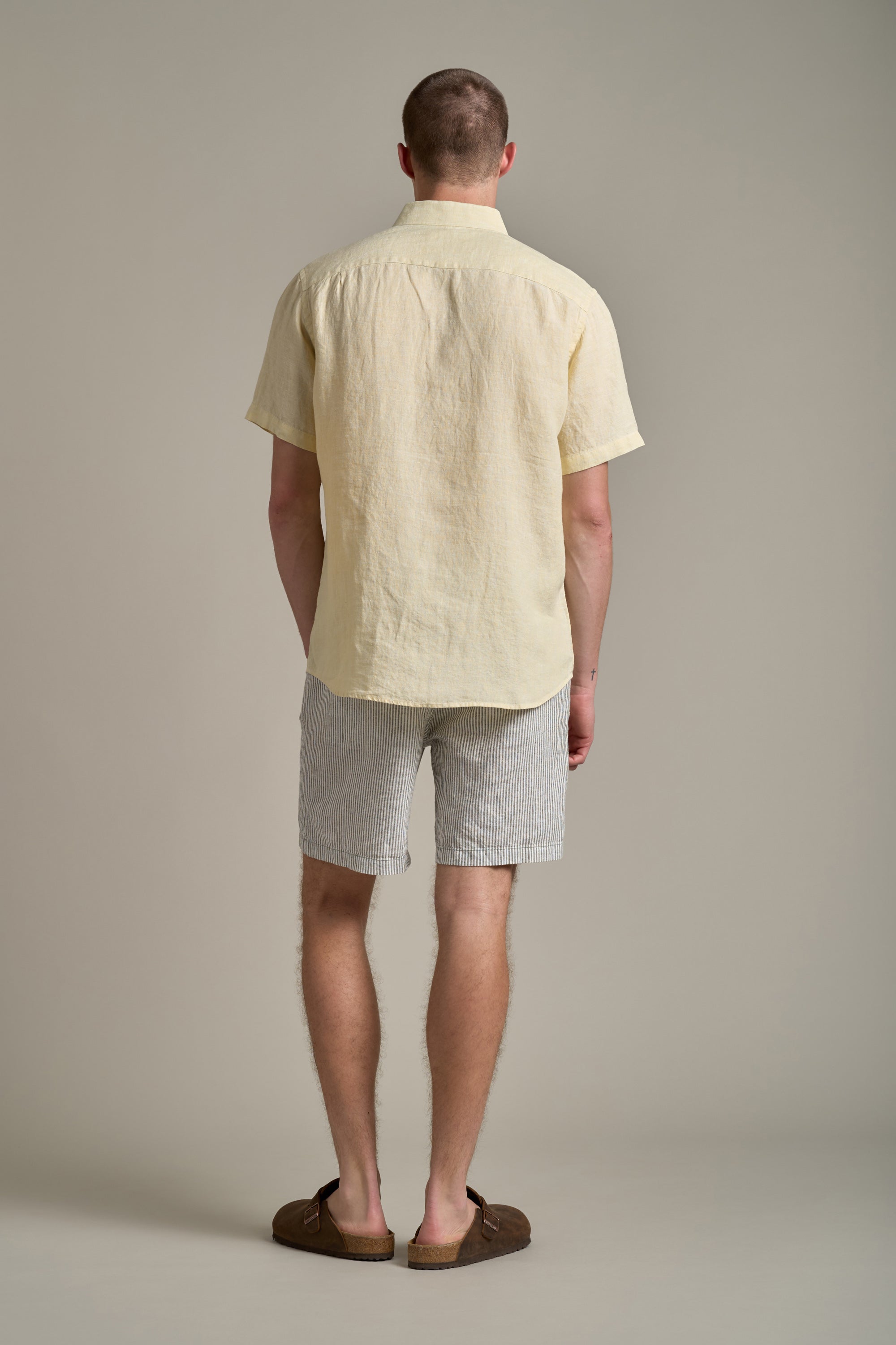 A man faces away from the camera wearing the Barkers Gentry Linen Shirt in pale yellow, paired with light gray striped shorts and brown shoes—an effortlessly cool addition to any summer wardrobe, set against a plain light background.