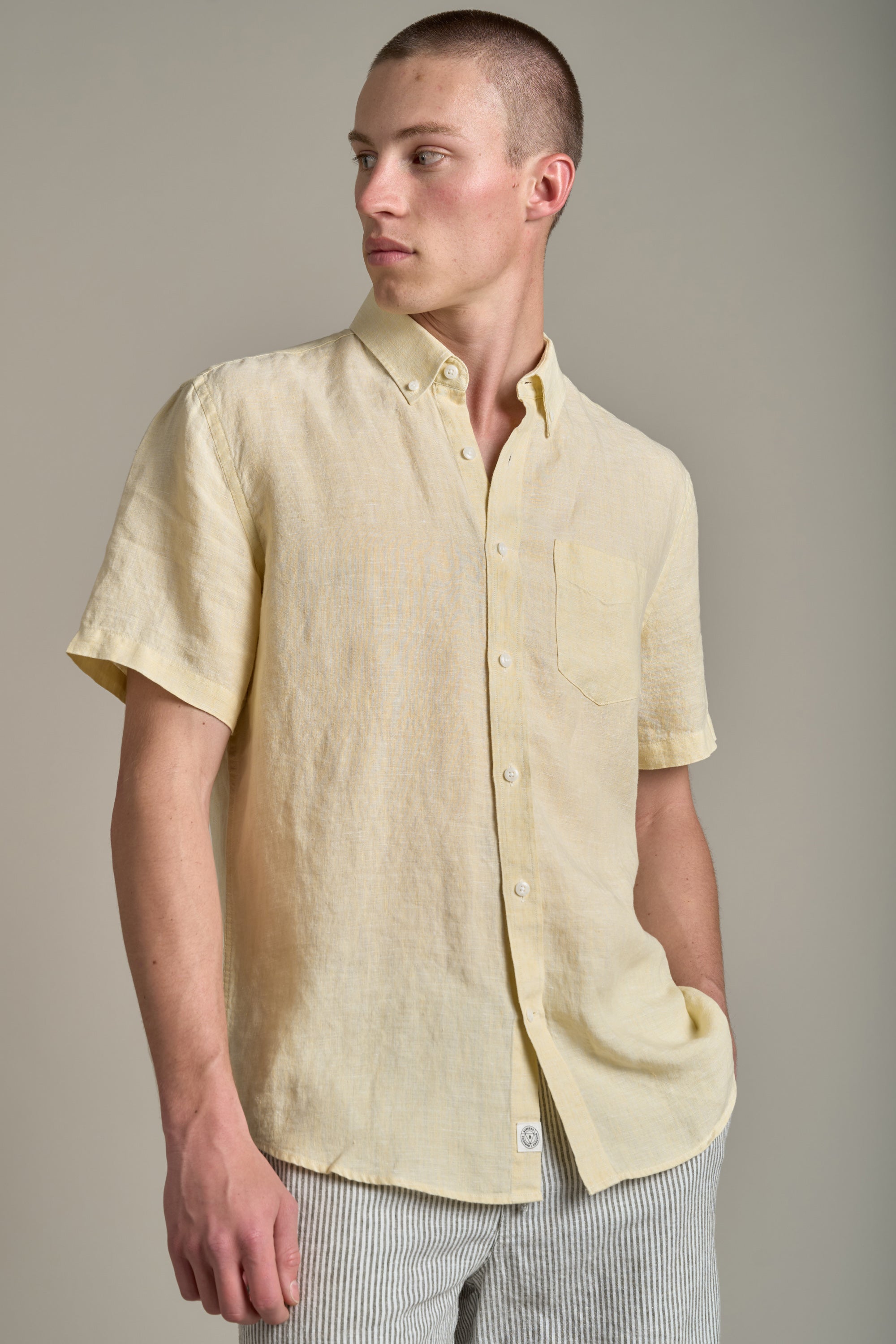 A man wears the Barkers Gentry Linen Shirt in light yellow with a chest pocket, paired with light gray striped pants—an ideal look for your summer wardrobe.