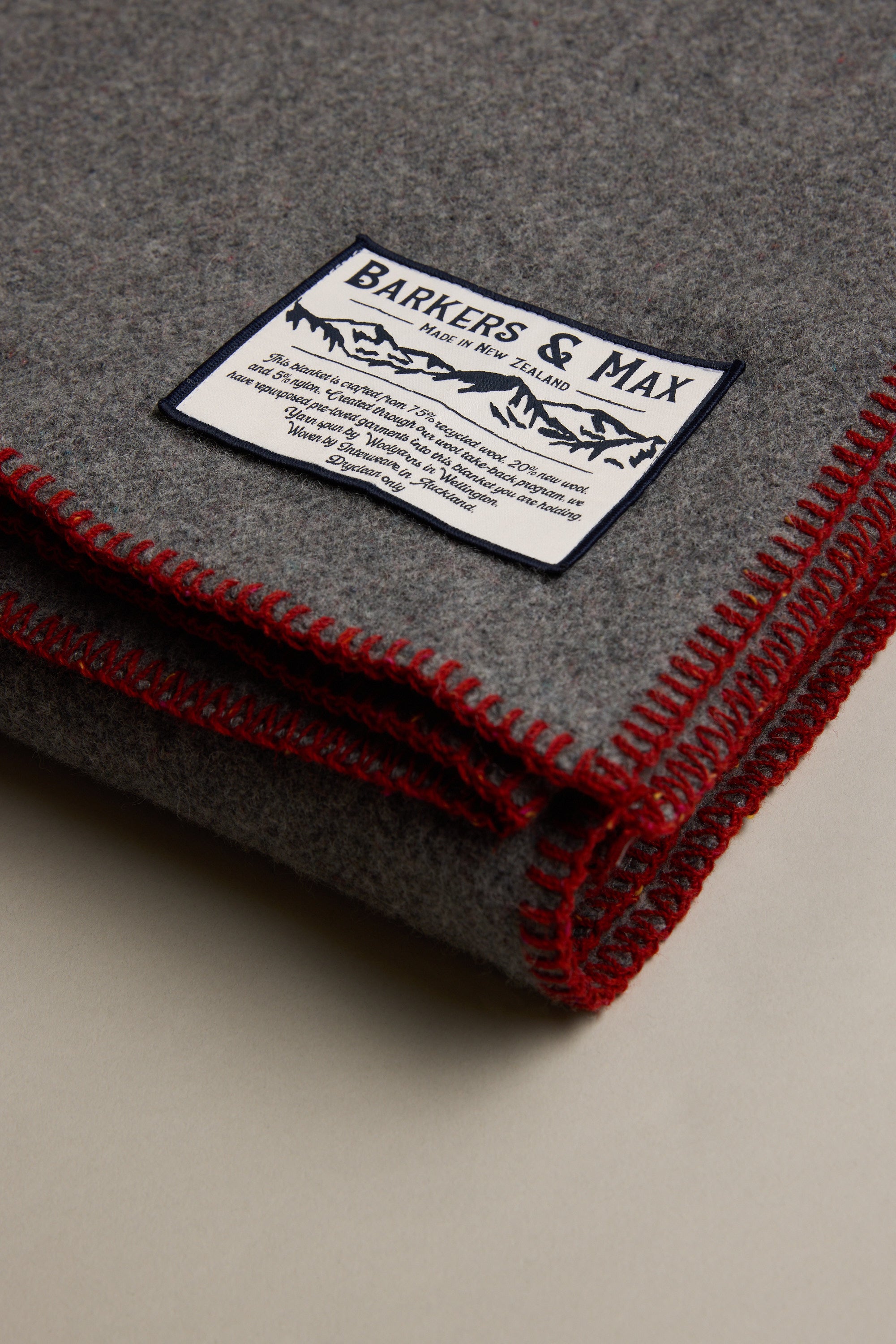 The Wool Take Back Blanket by Other is a folded grey recycled wool blanket with red edge stitching and a rectangular Barkers & Max label sewn on top.