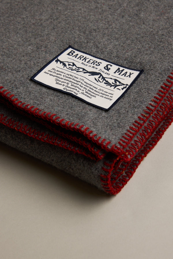 The Wool Take Back Blanket by Other is a folded grey recycled wool blanket with red edge stitching and a rectangular Barkers & Max label sewn on top.