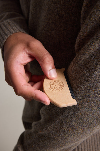 A hand uses the Barkers Sweater Comb on a brown wool sweater sleeve, gently removing pills and lint from the knitwear. The comb features an engraved Barkers logo and text.