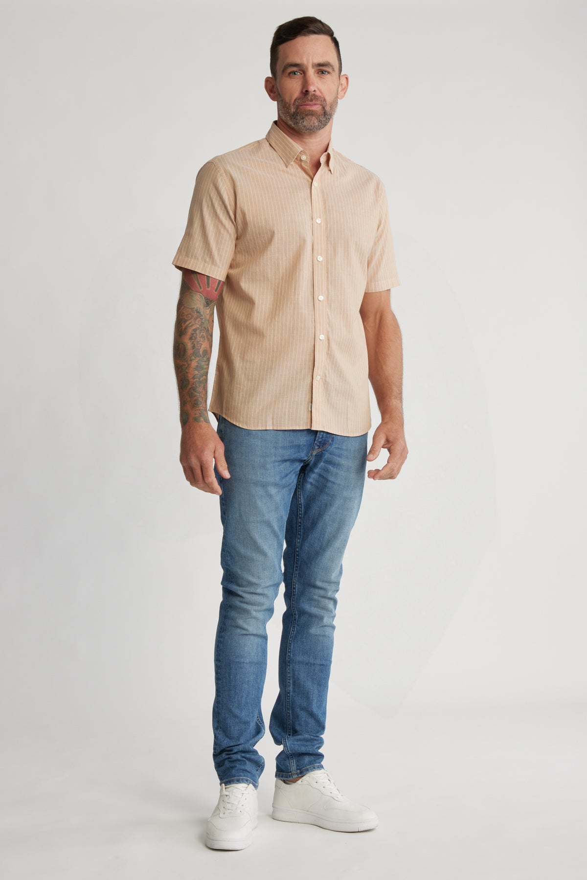 A man with short hair, a beard, and a tattooed arm stands against a plain background wearing the Barkers Burnham Stripe Shirt, blue jeans, and white sneakers.