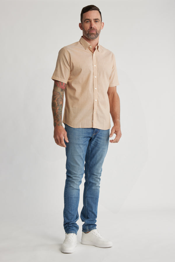 A man with short hair, a beard, and a tattooed arm stands against a plain background wearing the Barkers Burnham Stripe Shirt, blue jeans, and white sneakers.