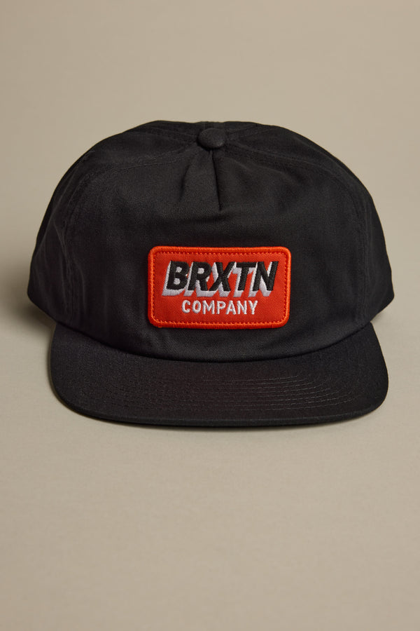 The Brixton Payment MP Snapback, a black 5-panel hat with a flat brim, features a red and white BRXTN COMPANY patch on the front. It is shown against a neutral beige background.