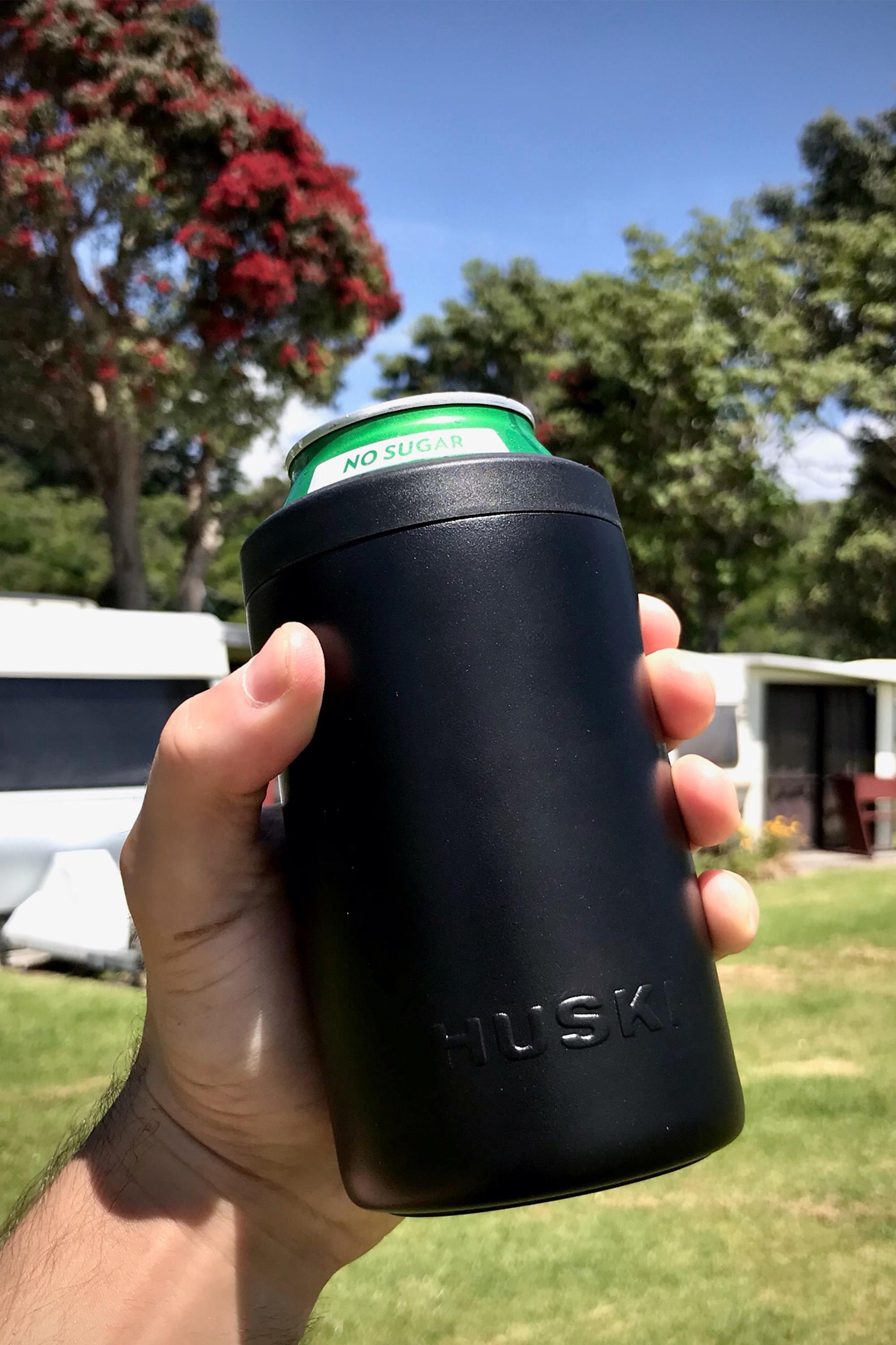 A hand holds a black Huski Beer Cooler by Huski with a green-labeled can outdoors, with trees, grass, a caravan, and a building in the sunny background.