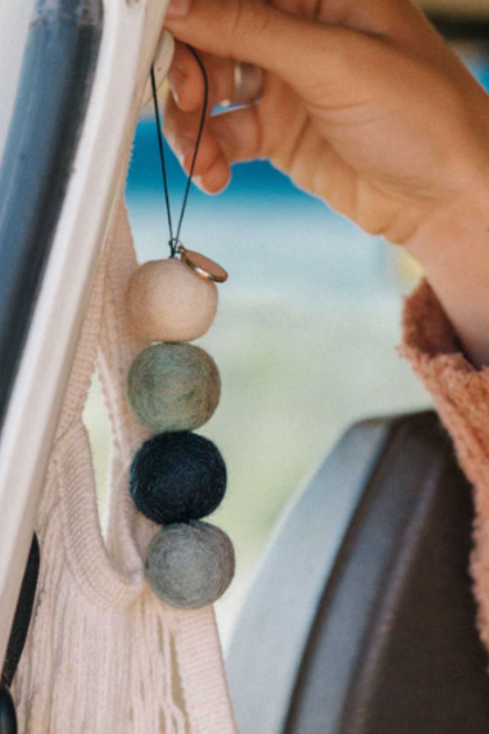 A hand hangs Other’s Smelly Balls: Cove / Coastal Drift—a string of four felted wool balls in cream, green, blue, and gray—from a car window frame with a blurred background and partial macrame decoration visible.
