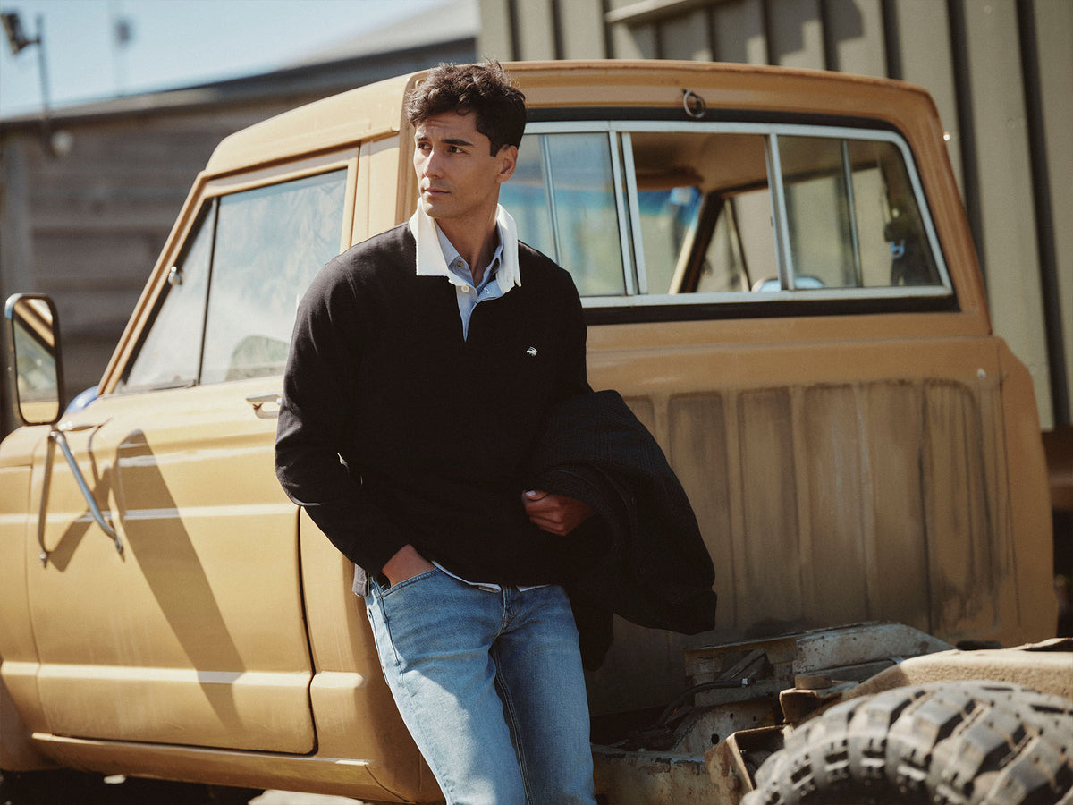 Man in black sweater and jeans leans against a vintage yellow NZ truck outdoors.