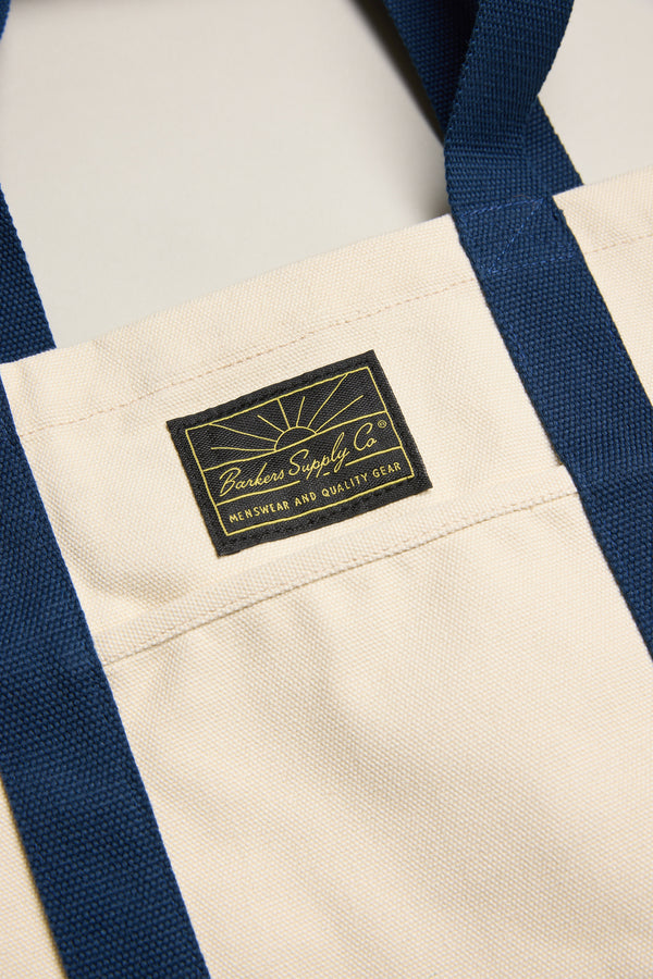 Close-up of a durable Barkers Supply Co. Canvas Tote Bag with dark blue handles and a black and yellow Barkers label stitched on this versatile travel tote.