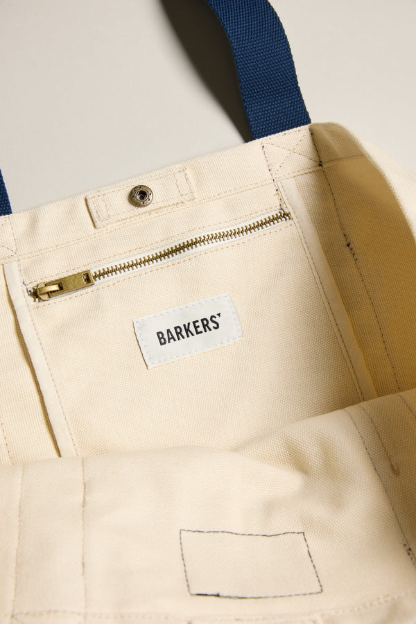 Close-up of the Barkers Supply Co. Canvas Tote Bag by Barkers, showing its durable canvas interior, zipped pocket, snap button, blue handles, and a white label with the Barkers logo.